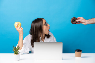 Hand offering donut to hispanic young woman holding fruit isolated on blue background. Food...