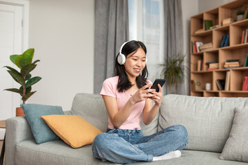 Obraz premium Online chat in social networks. Happy young korean lady in headphones typing on smartphone, resting on couch