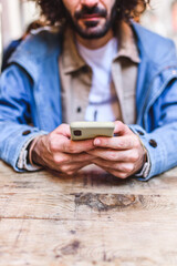 Soft focus of crop bearded man in casual clothes sending text message on mobile phone while sitting at lumber table in restaurant