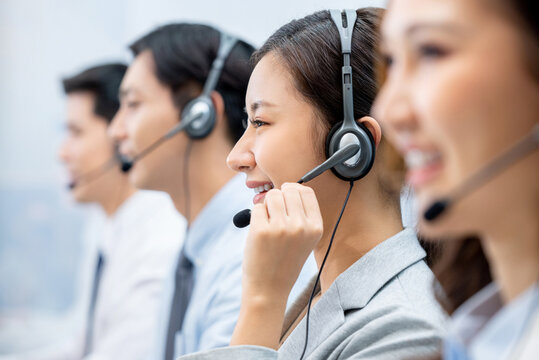 Side view of positive Asian woman telemarketer wearing headset with microphone working with colleagues in call center office