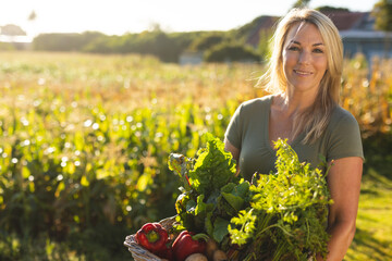 Portrait of smiling caucasian mid adult woman carrying vegetables against crops in farm on sunny day