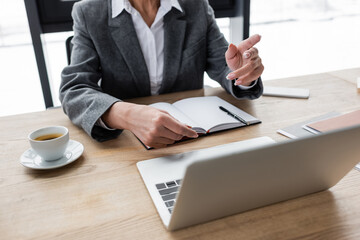 cropped view of banker pointing with finger during video call on laptop near notebook and coffee cup.