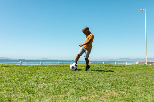 Full length of african american boy kicking soccer ball on grass against blue sky with copy space
