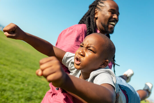 African american father carrying son while boy imitating as superhero flying on sunny day