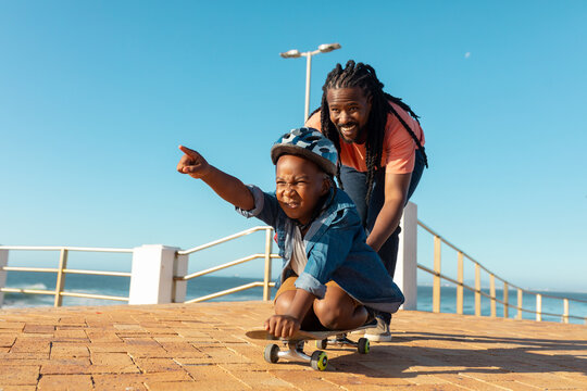 African american father pushing son pointing on skateboard at promenade against blue sky