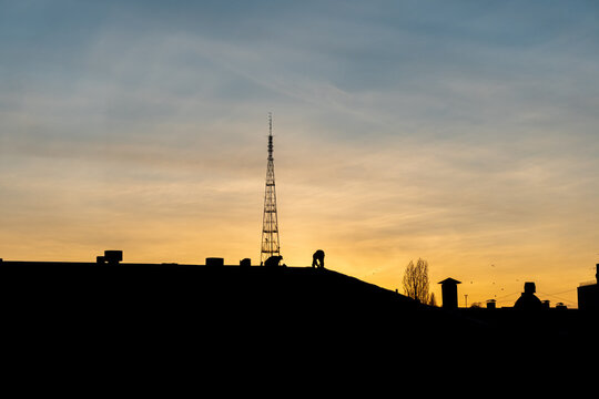 A Silhouette Of Two Men Working On A Roof Of A Building In The City At A Sunset Time. Golden Hour, Big Tower And Residential Buildings.