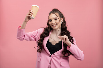 Portrait of an elegantly dressed woman in a jacket holding a takeaway cup wireless headphones girl dances happily against a pink studio background.