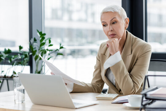senior financier holding documents while sitting near laptop.