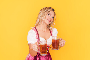 Woman in festive dress holding two mugs with beer in front of yellow background.