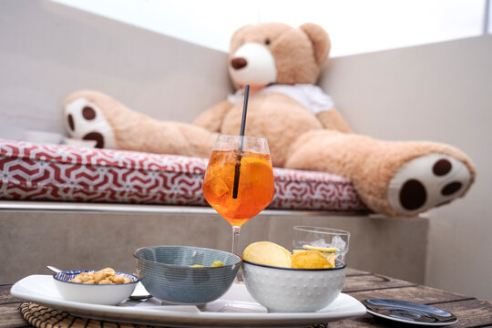 Funny image of a big teddy bear drinking spritz aperitif cocktail drink sitting on the sofa - Party night concept - Focus on red cocktail