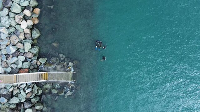 A group of scuba divers gather above the water near a set of stairs leading to the ocean. High drone view