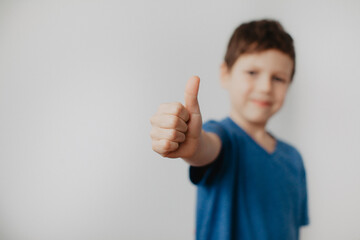 A preschool boy in a blue T-shirt on a light background shows a thumbs up