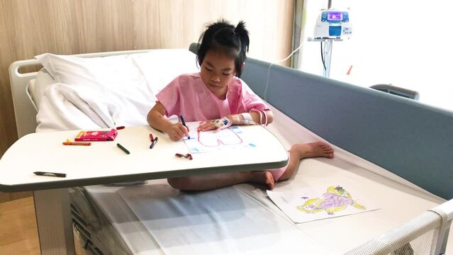 Asian patient child girl sitting on hospital bed, the 6 years old child is drawing as relax time in hospital's room, hand with infusion needle and IV tube for intravenous infusion. Light from window.