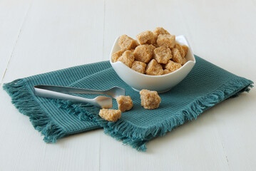 Cube-shaped cane sugar in wooden bowl on a turquoise napkin with silver sugar tongs. Front closeup view, white wooden table, no people.