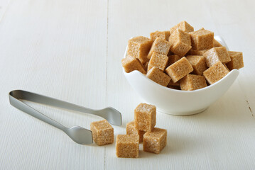 Cube-shaped cane sugar in porcelain bowl on a white wooden table with silver sugar tongs. Front closeup view, no people.