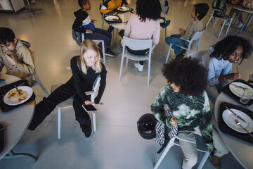 Girl sharing smart phone with friend sitting at table during lunch break in cafeteria