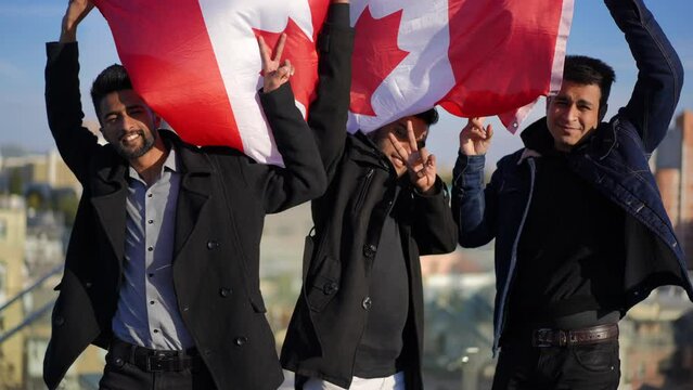 Three positive men with Canadian flag looking at camera gesturing peace standing on rooftop. Portrait of confident Middle Eastern immigrants posing in sunshine outdoors. Lifestyle concept
