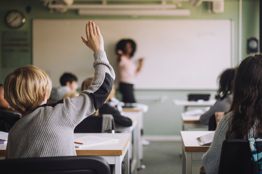 Rear view of student raising hand while teacher teaching in classroom