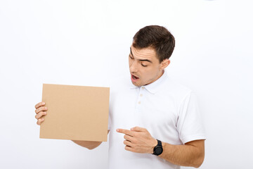 portrait of surprised brunette man with short hair and open mouth in white T-shirt on white background points finger at empty paperboard for advertising, inscriptions, copy space, template