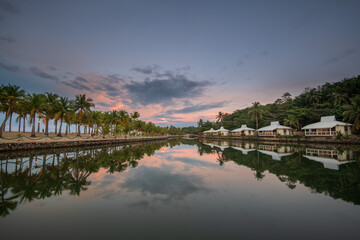 A lake with reflection of white bungalows in a row on the coastline against palm trees and blue sky