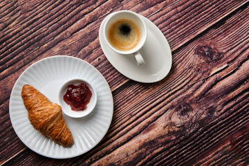 A Croissant, strawberry jam & espresso coffee on a white textured round plate on a wood background taken from above, a breakfast favourite worldwide.