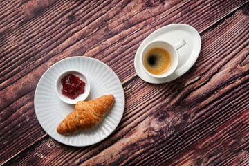 A Croissant, strawberry jam & espresso coffee on a white textured round plate on a wood background taken from above, a breakfast favourite worldwide.