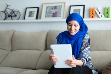 Smiling young Muslim woman using a digital tablet at home. Technology and lifestyle. Space for text.