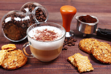 Cup of cappuccino, crackers, cookies, holder with ground coffee, tamper and coffee beans on wooden table.