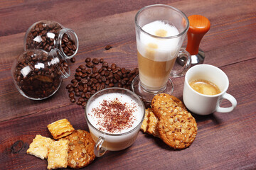Breakfast with espresso, latte and cappuccino, crackers, holder with ground coffee, tamper and coffee beans on wooden table.