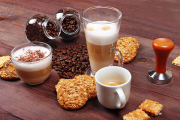 Still life with espresso, latte and cappuccino, crackers, holder with ground coffee, tamper and coffee beans on wooden table.