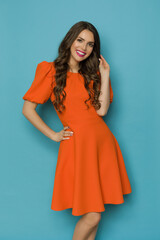 Beautiful smiling young woman in orange cocktail mini dress is posing with hand on hip.