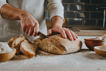 Young man in apron cutting homemade bread - obrazy, fototapety, plakaty