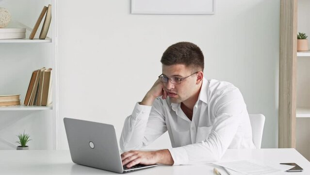 Bored man. Clueless work. Silly tasks. Disinterested guy in white shirt looking laptop with dull emotion sitting desk leaning hand in light room interior.