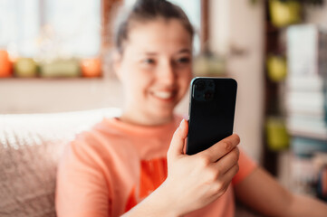 Fototapeta na wymiar Teen girl checking social media holding smartphone at home. Smiling young woman using mobile phone app playing game, shopping online.