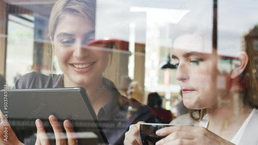 Poster Two businesswomen planning during a meeting in a cafe while drinking coffee and using a wireless tablet. Two smiling businesswomen collaborate and talk while scrolling online on a digital tablet.