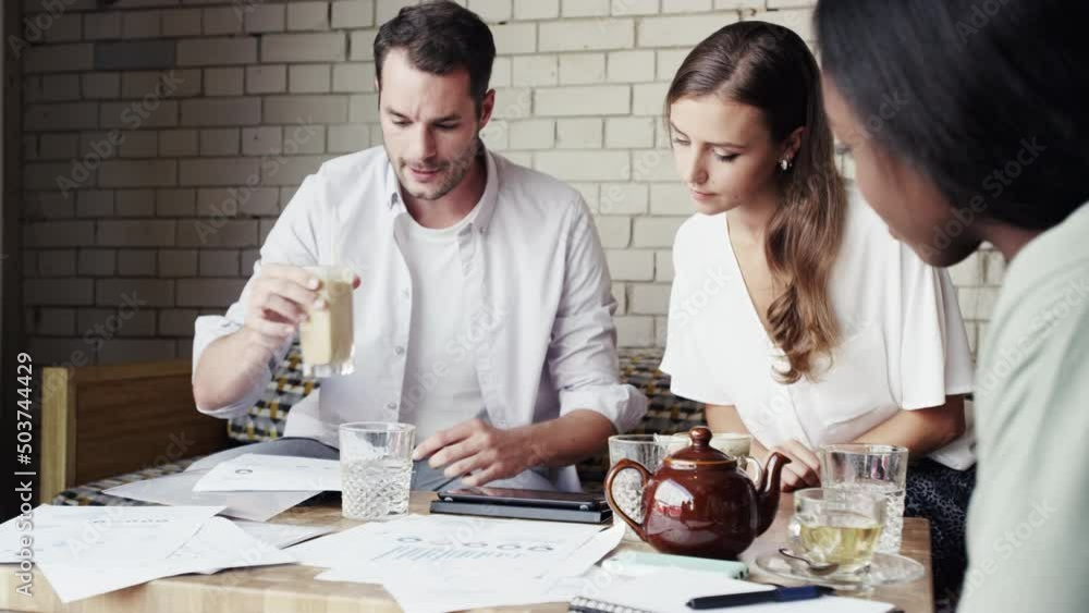 Poster a young businessman drinking coffee during a meeting looking over architectural drawings with collea