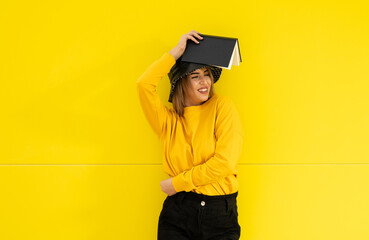 A young caucasian woman in a black hat holding a book over a yellow colored wall
