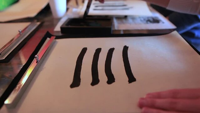 Close-up of a hand and a calligraphy brush. A person learns to hold a brush with black ink at the right angle and draw elements of hieroglyphs on paper. Drawing vertical lines.