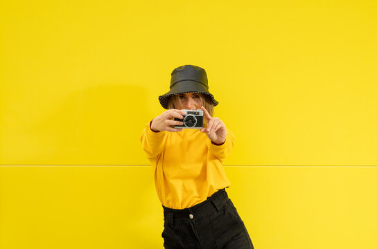 A young caucasian woman in a black hat holding a vintage photo camera over a yellow colored wall
