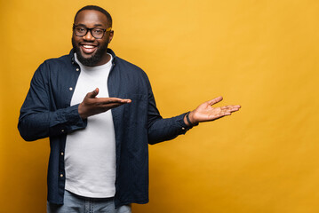 Smiling cheerful young African American man points both hands aside looking at the camera with...