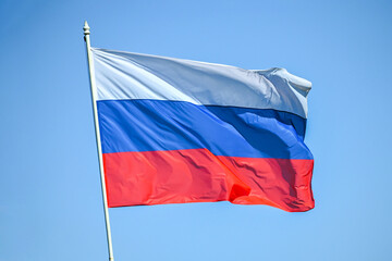 Flagpole with the Russian tricolor flag fluttering in the wind against a blue sky. The flag is the national symbol of the country. 