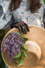 Woman's hand in vintage black lace glove with ring holds a straw hat with branch of lilac flowers. Cottagecore concept.