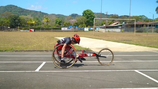 A kid with disability driving a handbike at a paracycling race, striving to win. Handheld slow motion tracking shot (side of the bike running on the track).