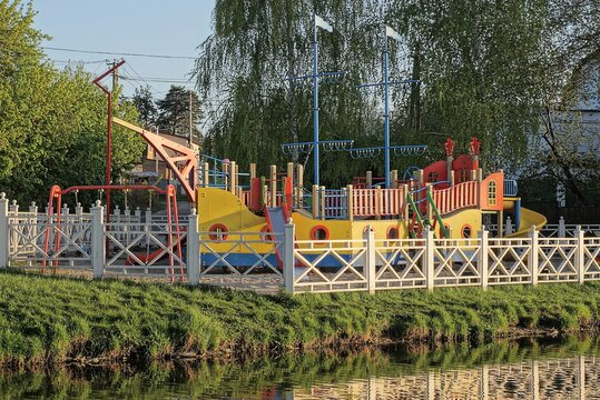 Playground With A Model Of A Large Colored Ship Behind A White Wooden Fence In Green Grass On The Shore Near The Water Of A Reservoir