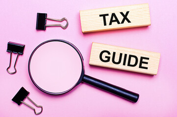 On a pink background, a magnifier, black paper clips and wooden blocks with the text TAX GUIDE. Business concept