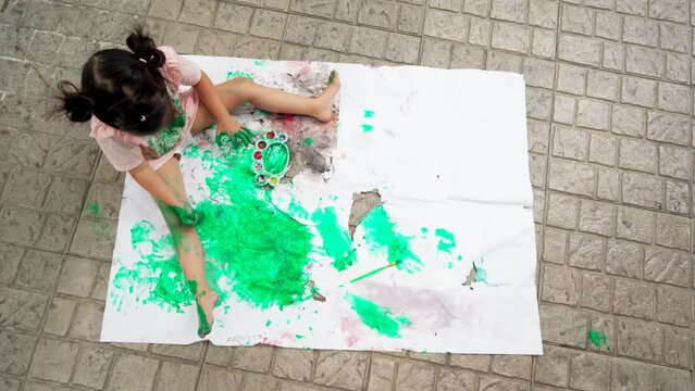 Asian child girl sitting on rip canvas on tile floor in the garden.Painted green color on hands so dirty, leave a mark on the white paper. Children's creativity. Overhead shot of painting process. 