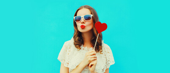 Portrait beautiful young woman with red heart shaped lollipop blowing lips sending sweet air kiss on blue background