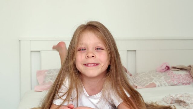 portrait of a smiling little girl. A beautiful, sweet little girl with blue eyes and long hair lies on her stomach in bed and smiles. The child is naughty and indulges. Children's pranks and games in 