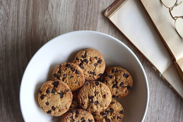 Plate of chocolate chip cookies, open books and reading glasses on wooden table. Flat lay.
