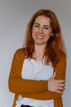 Happy middle aged ginger haired woman in her 40s over white wall. Employee in casual outfit looking at the camera. Female with long hair, cheerful freelancer business portrait. Copy space